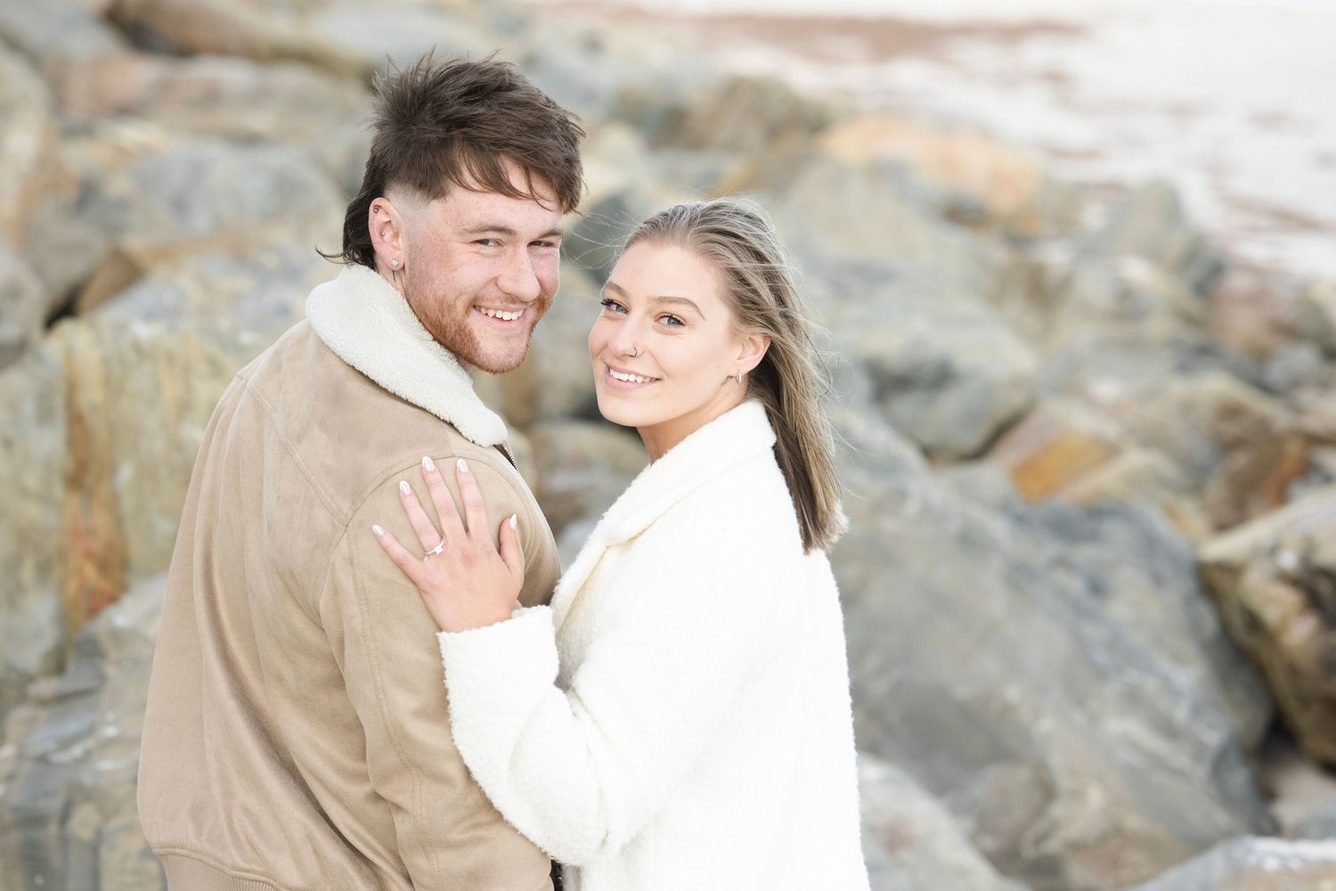 Happy couple embracing outdoors near rocky shore, wearing cozy winter jackets, smiling warmly at camera.