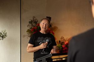 Person wearing a black AC/DC t-shirt stands smiling in front of a wall decorated with red flowers.