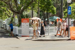 People walking along a tree-lined urban street with white fencing and orange barriers during a sunny day.