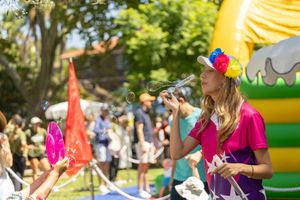 A person in a pink shirt and colorful headpiece speaks into a microphone at an outdoor event with a bouncy castle.
