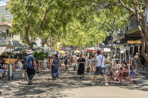 Tree-lined pedestrian street with outdoor dining and shops in a bustling urban neighborhood on a sunny day.