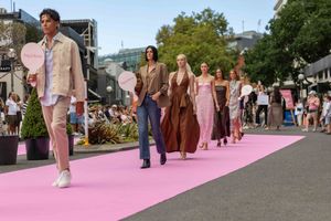 Models walk down a bright pink runway during an outdoor fashion show presentation on a city street.