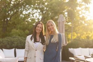Two women stand together outdoors at sunset with white patio furniture in the background.