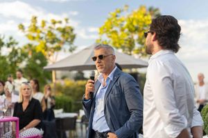 People gathered outdoors at a casual social event on a sunny day with trees and buildings in the background.