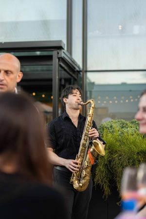 A musician performs on saxophone at an outdoor venue with string lights and greenery in the background.