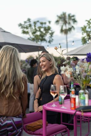 Group of friends enjoying drinks and conversation at an outdoor patio with pink tables and palm trees in background.