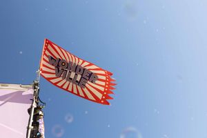 An orange and black striped flag waves against a bright blue sky with lens flare effects.