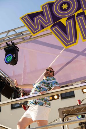 A performer in a Hawaiian shirt and white shorts stands on stage under a festival banner with stage lighting.