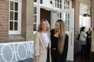 Two people in beige and black outfits stand and smile together against a brick wall exterior.
