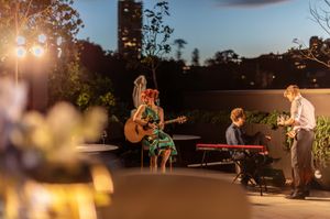 Musicians perform at an outdoor evening venue with acoustic guitar, keyboard and trumpet under warm lighting.