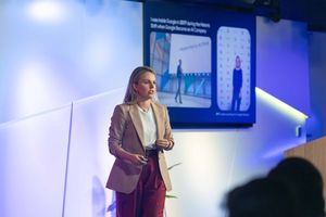 A speaker presents on stage at a business conference with blue lighting and presentation screens in the background.