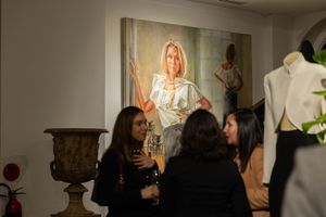 Two people chat in front of a large illuminated portrait painting hanging on a wall in an art gallery.