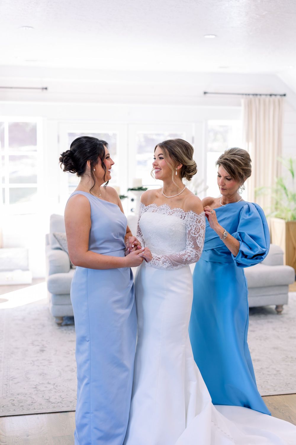 Wedding party in shades of blue dresses gather around bride in white lace gown indoors.