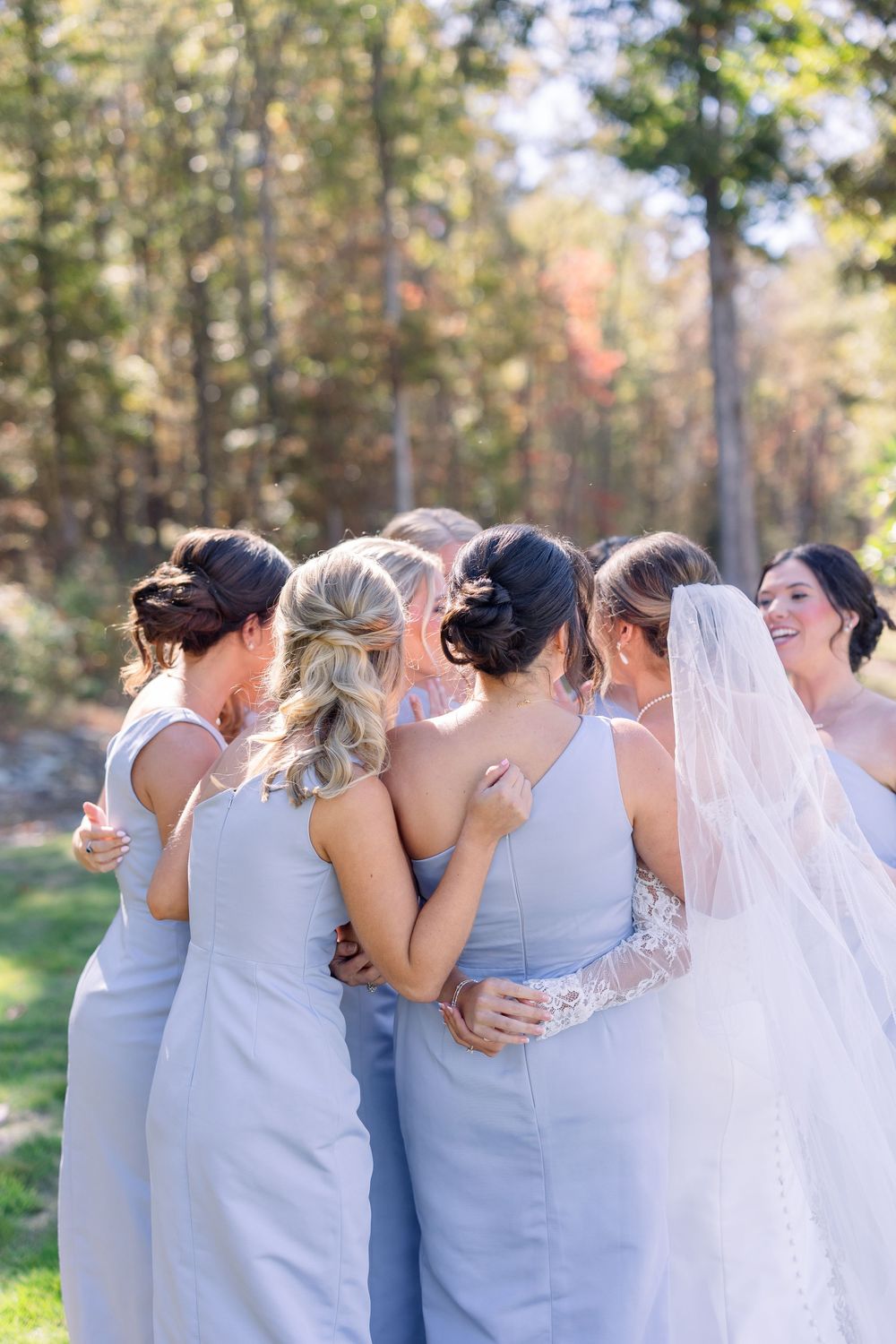 A group of bridesmaids in light blue dresses embrace together outdoors during a wedding photo session.