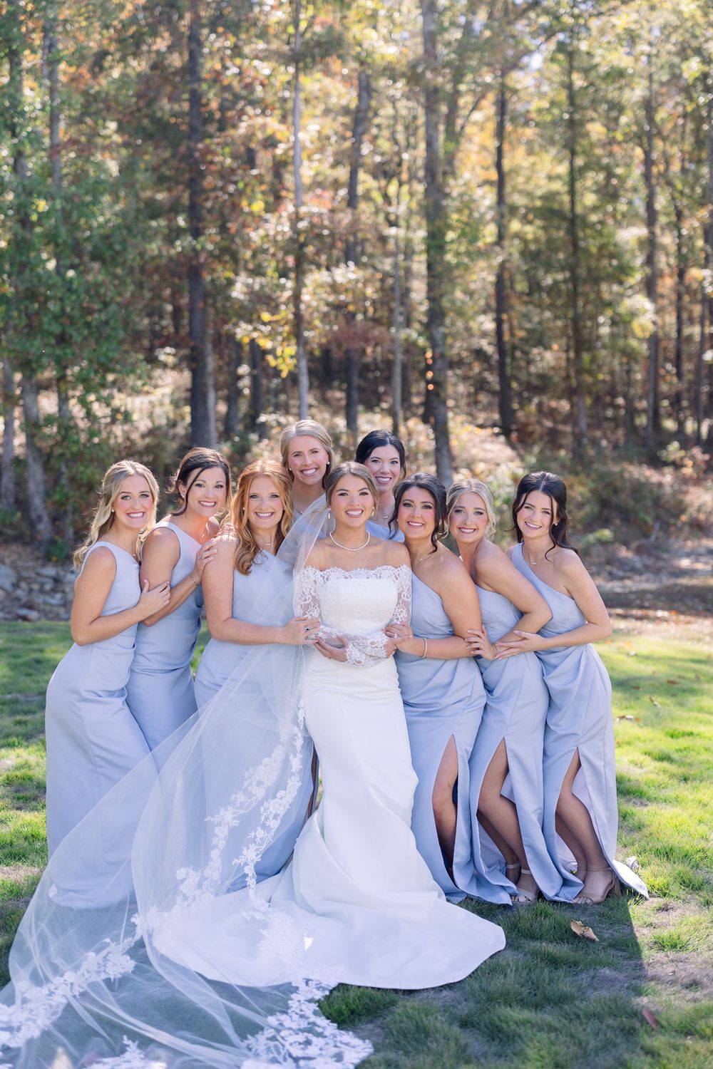 Bridesmaids in light blue dresses pose together on grass with autumn trees in background.