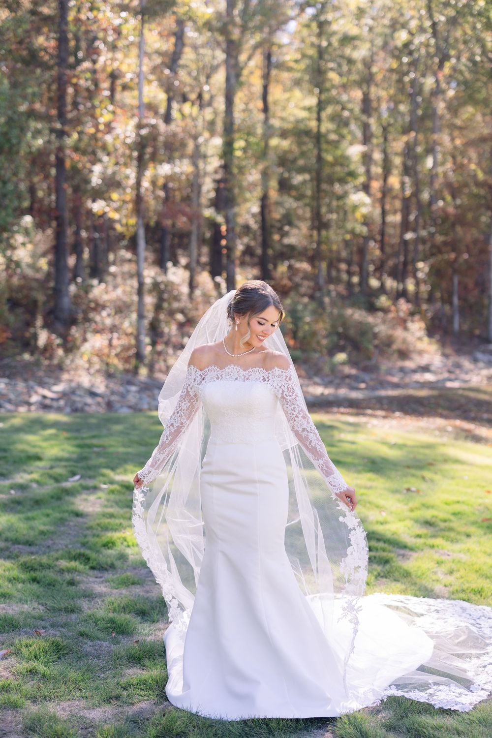 Elegant white lace wedding gown with long sleeves and flowing veil photographed in a sunlit autumn forest setting.