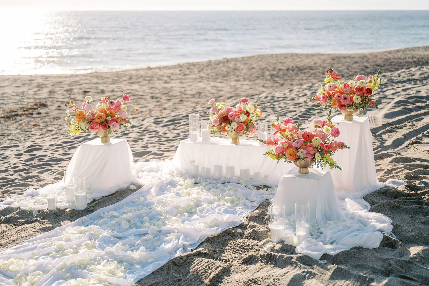 A romantic beach wedding setup with white tables and coral floral arrangements along the sandy shoreline at sunset.