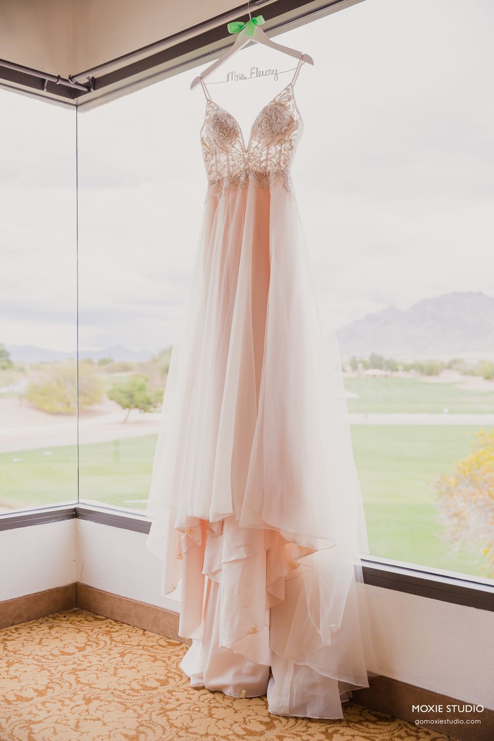 A blush pink wedding gown with lace bodice and flowing chiffon skirt hangs by a window overlooking mountain scenery.