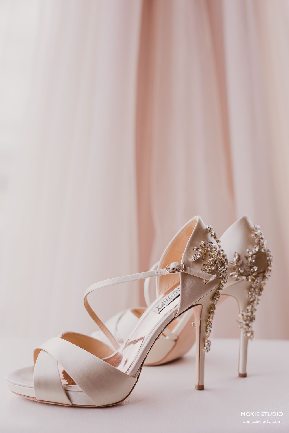 Elegant white satin bridal shoes with crystal embellished heels displayed against a soft pink backdrop.