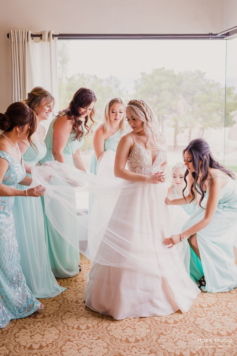 Bridesmaids in mint green dresses help the bride with her wedding gown in a sunlit room.