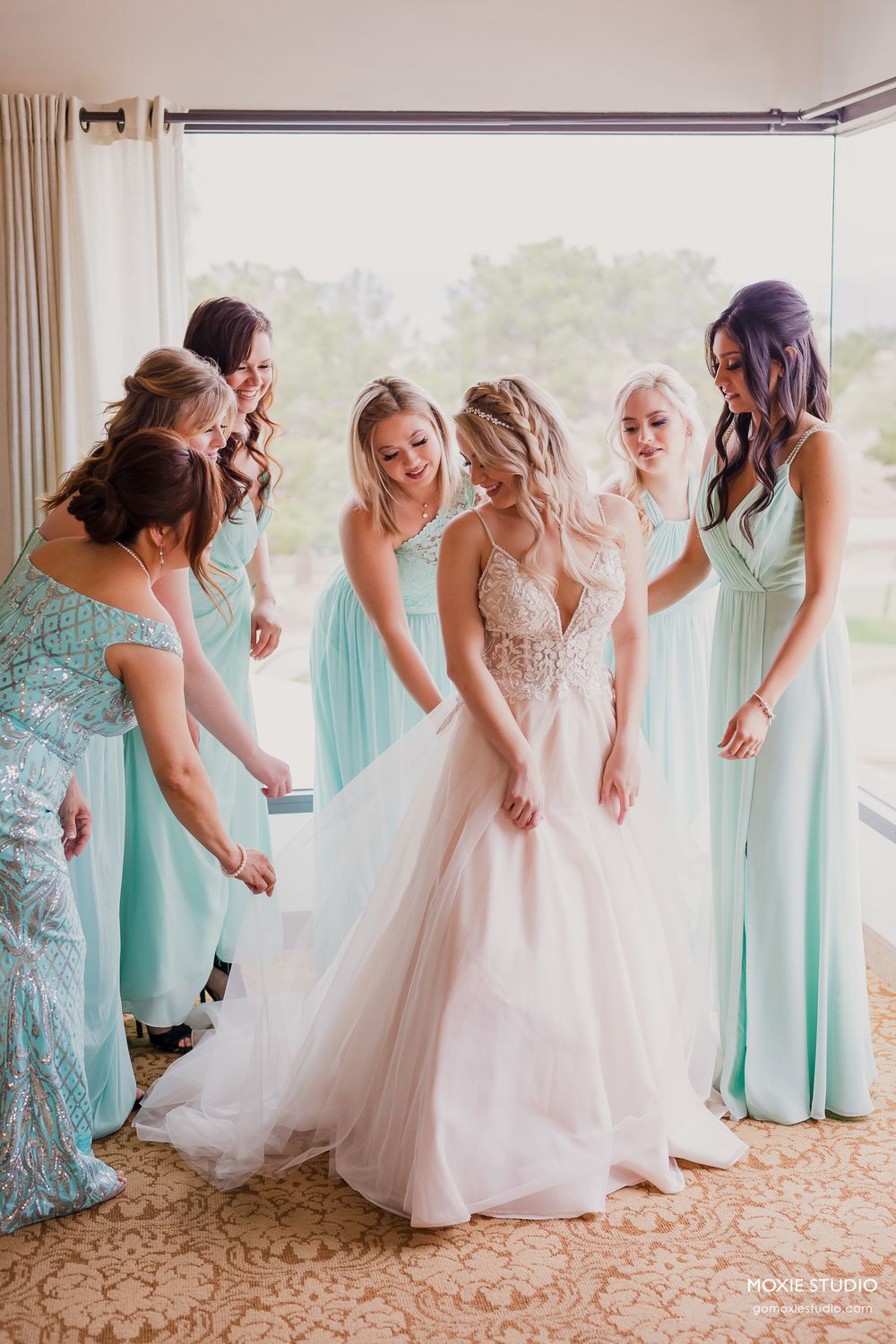 Bridesmaids in mint green dresses gather around bride in blush gown while helping with wedding preparations.