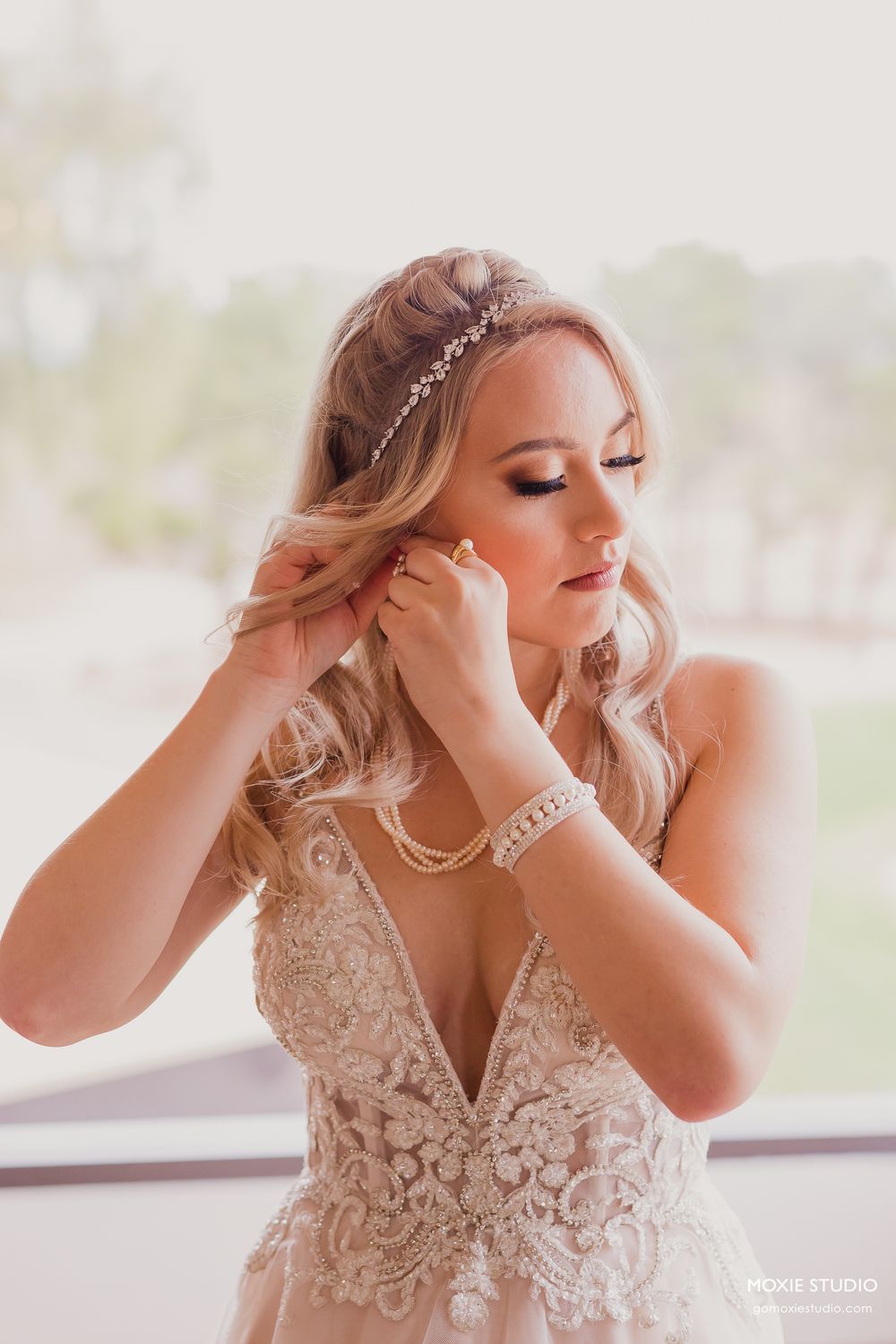 A bride wearing a delicate lace wedding gown and crystal headpiece puts on earrings in soft natural light.