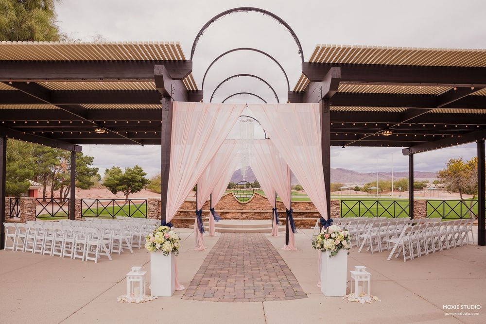 An outdoor wedding ceremony setup with white chairs and metal arches creating a tunnel effect leading to the altar.