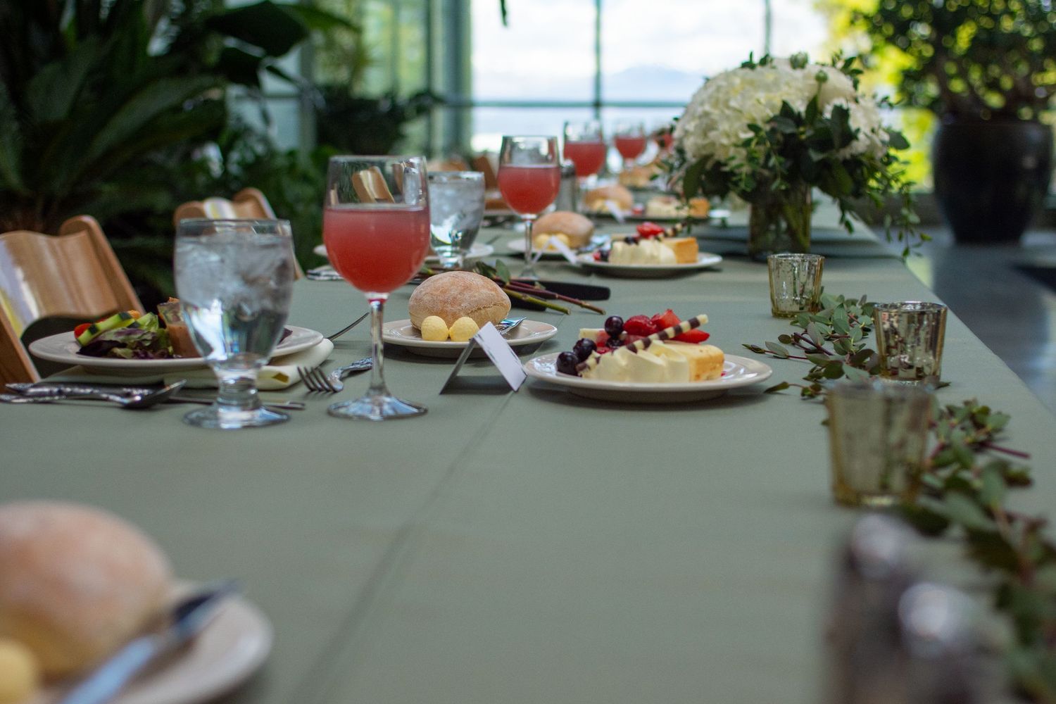 An elegant outdoor dining table set with plates and glassware surrounded by lush greenery and natural lighting.