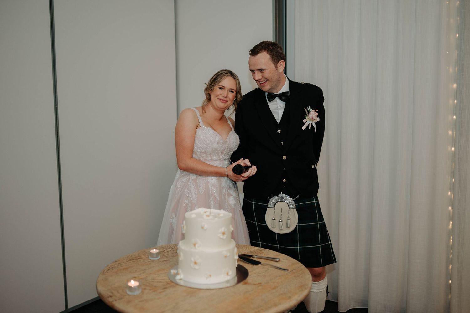 A couple cuts their white wedding cake together at their reception while wearing formal attire.