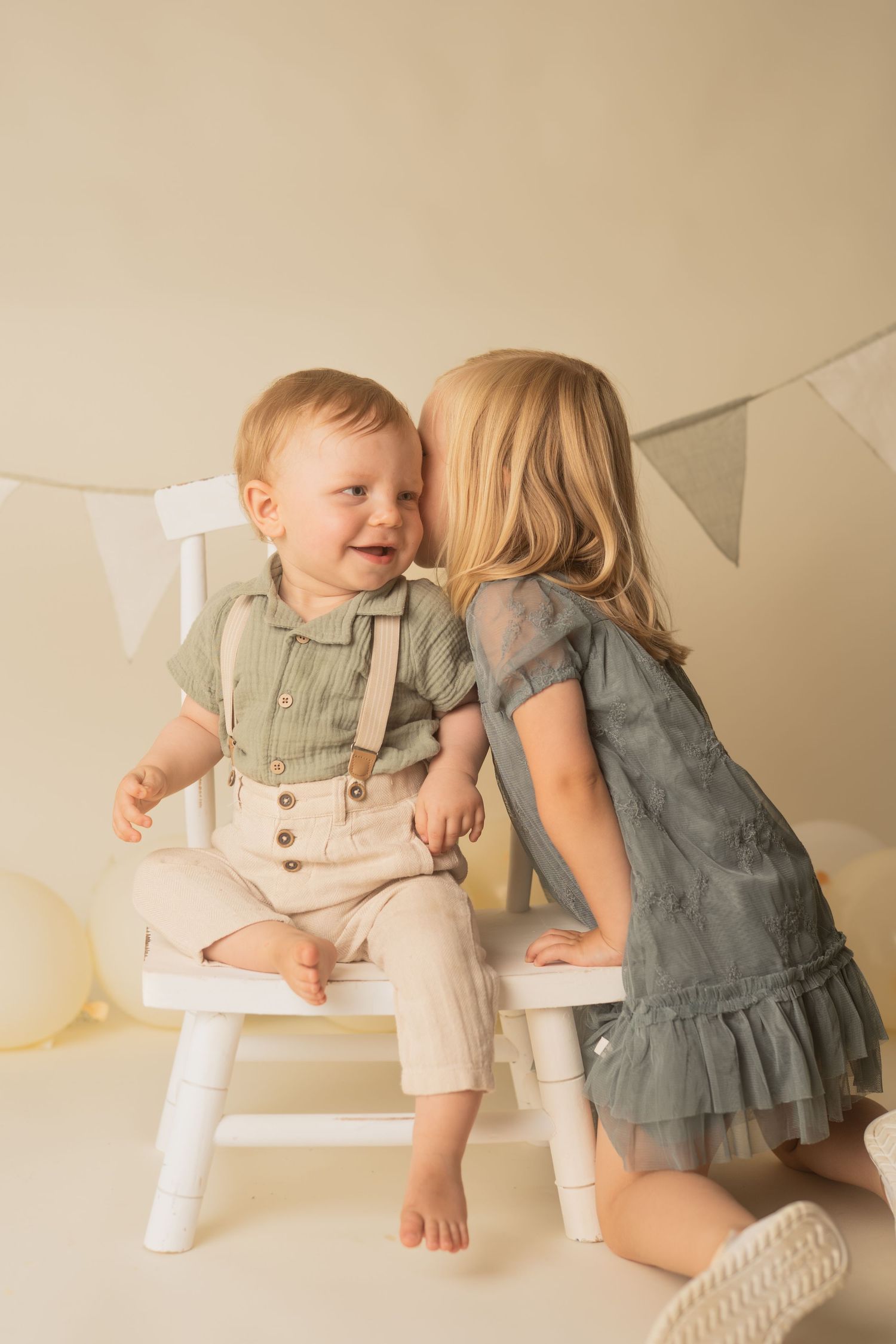 Sweet baby wearing beige linen overalls sits on floor with decorative bunting in background.