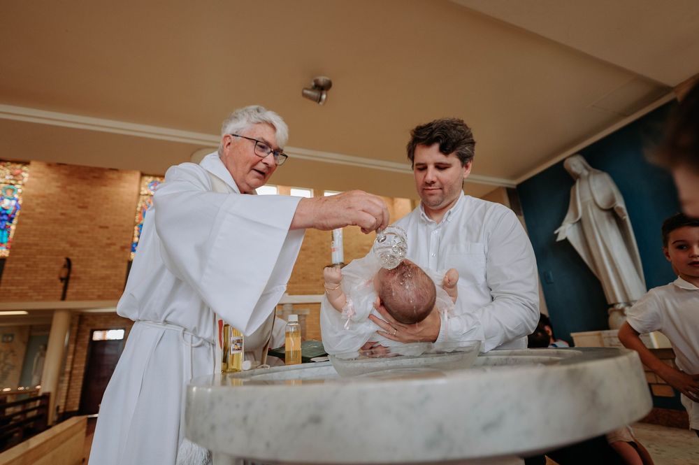 A baptism ceremony takes place at a church font with clergy and family members gathered around.