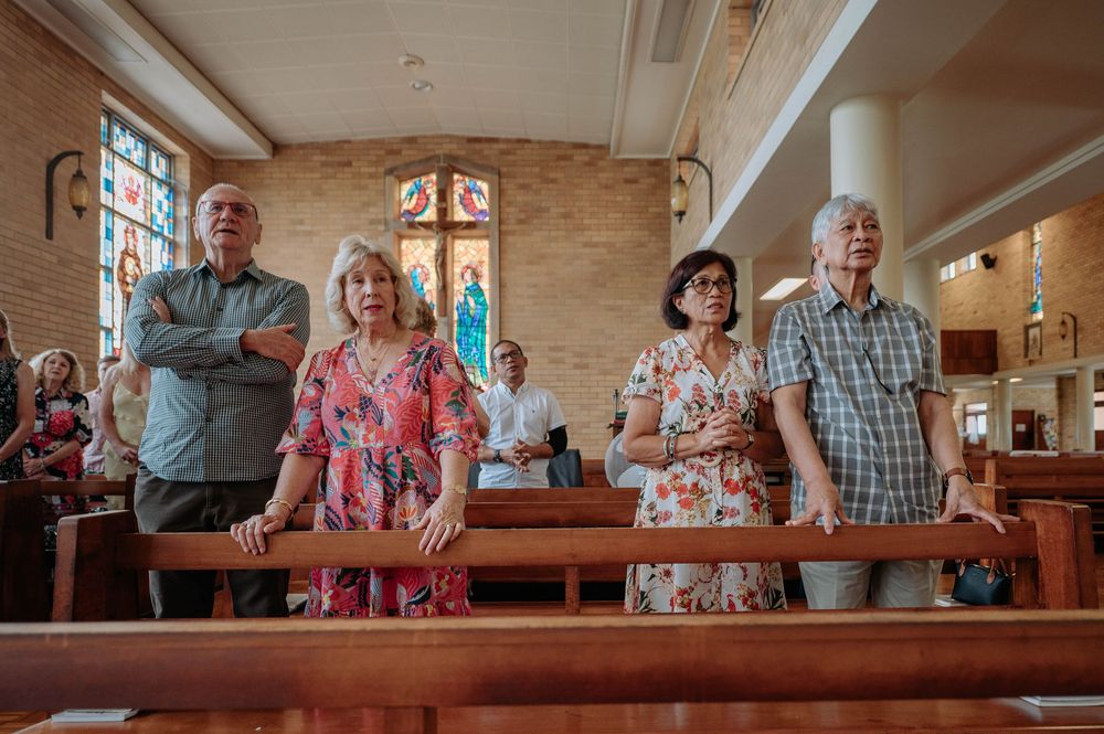 People standing in wooden church pews during a service with stained glass windows visible in the background.