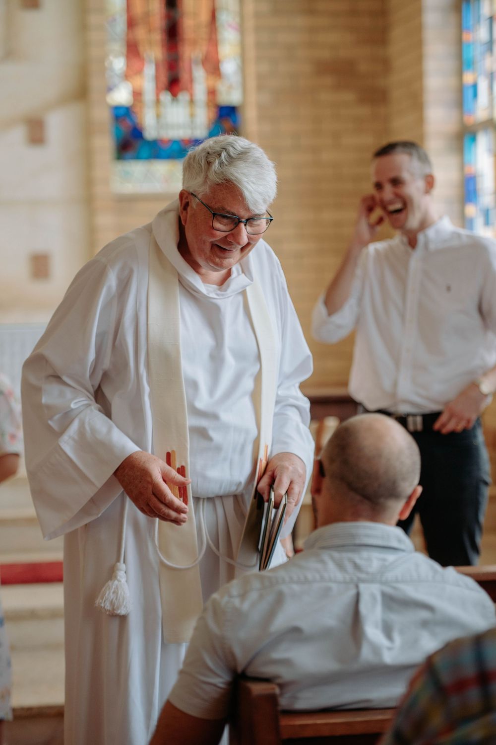 A clergy member in white robes shares a joyful moment during a church service with stained glass windows in the background.