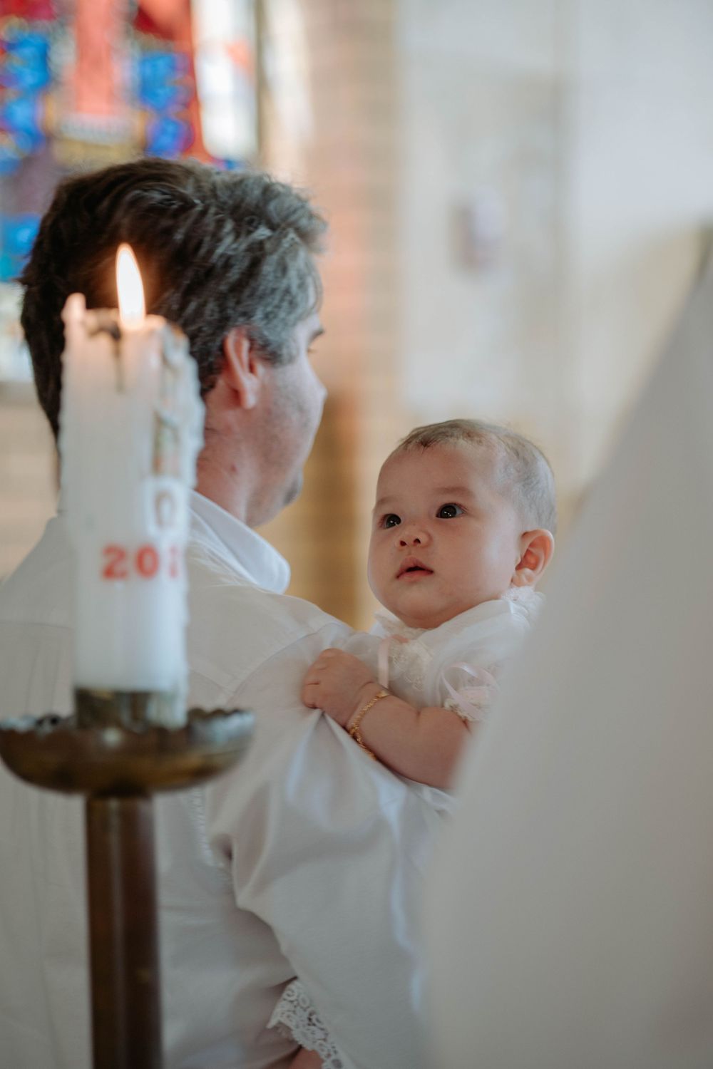 A small baby in white christening attire is held near a lit ceremonial candle during a baptism service.