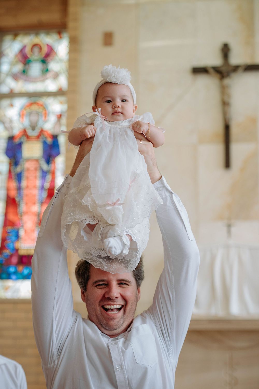 A series of four photos showing a baby in white christening dress being lifted up inside a church with stained glass windows.