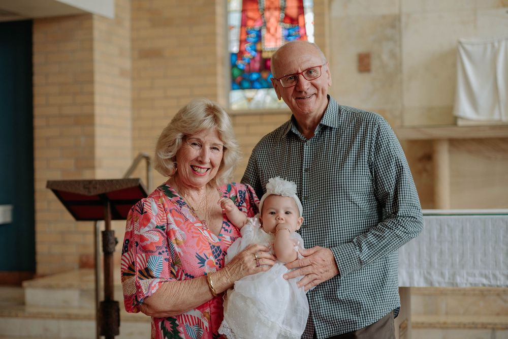 A couple holds a baby in white christening dress inside a church with stained glass windows.