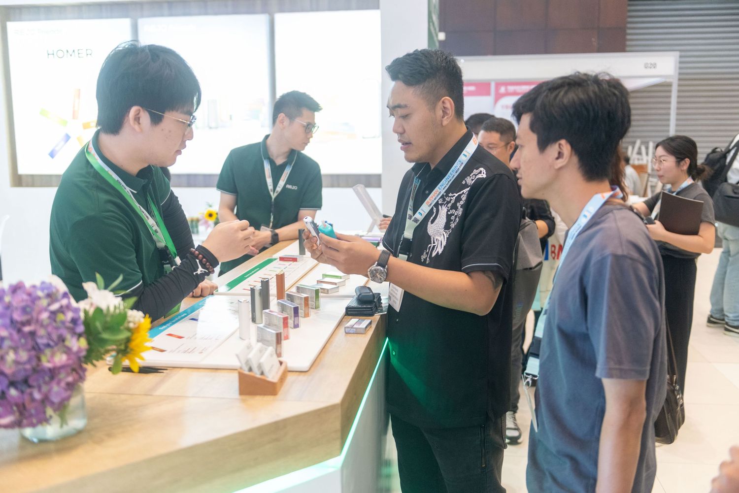 Customers interact with staff at a retail counter with product displays and purple flowers in the foreground.