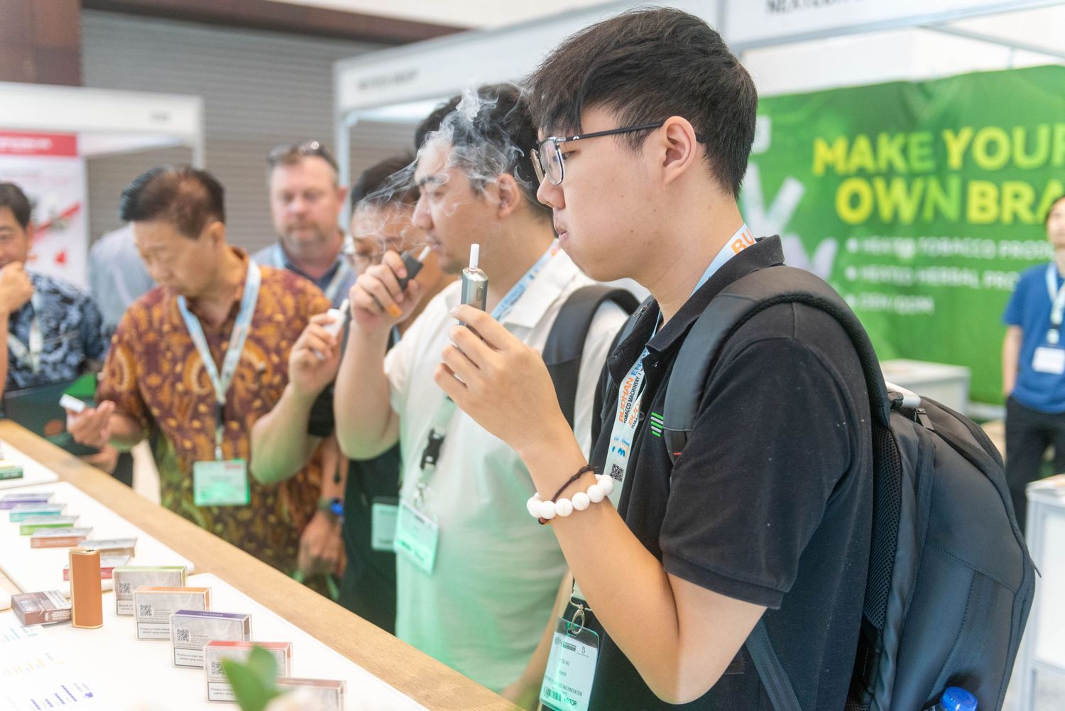 People gather at a business exhibition or trade show with display booths and promotional banners visible in the background.