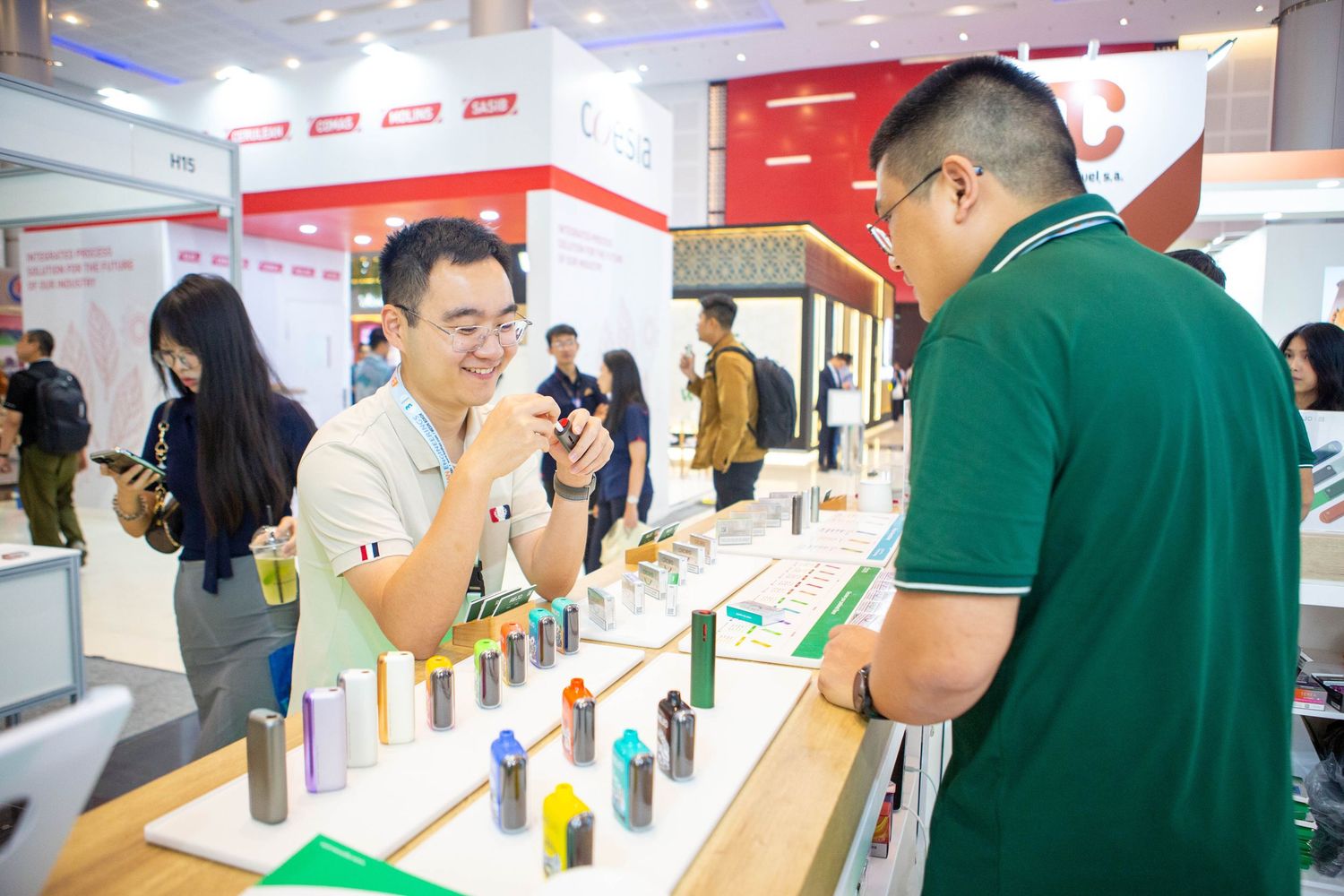 People browse and sample products at a trade show booth displaying various bottles and containers.