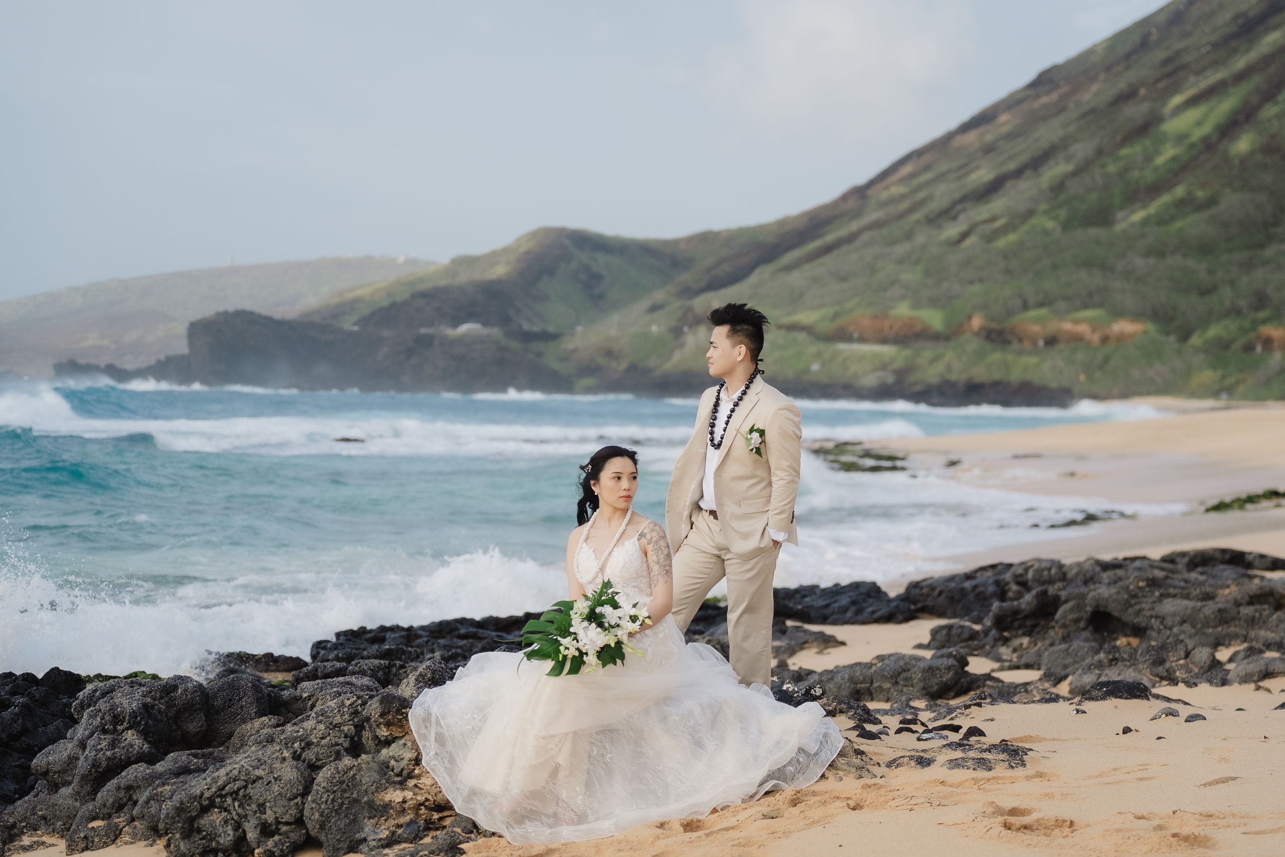 A wedding couple poses on a dramatic Hawaiian beach with crashing waves and volcanic rocks.