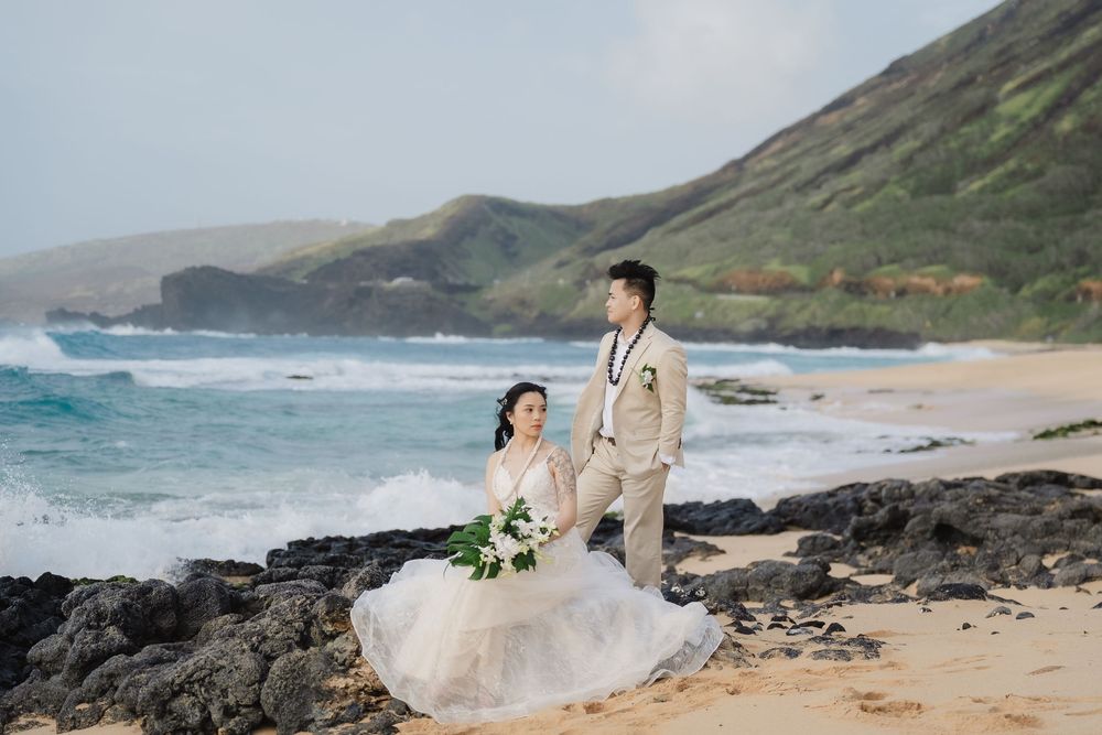 A wedding couple poses on a dramatic Hawaiian beach with crashing waves and volcanic rocks.