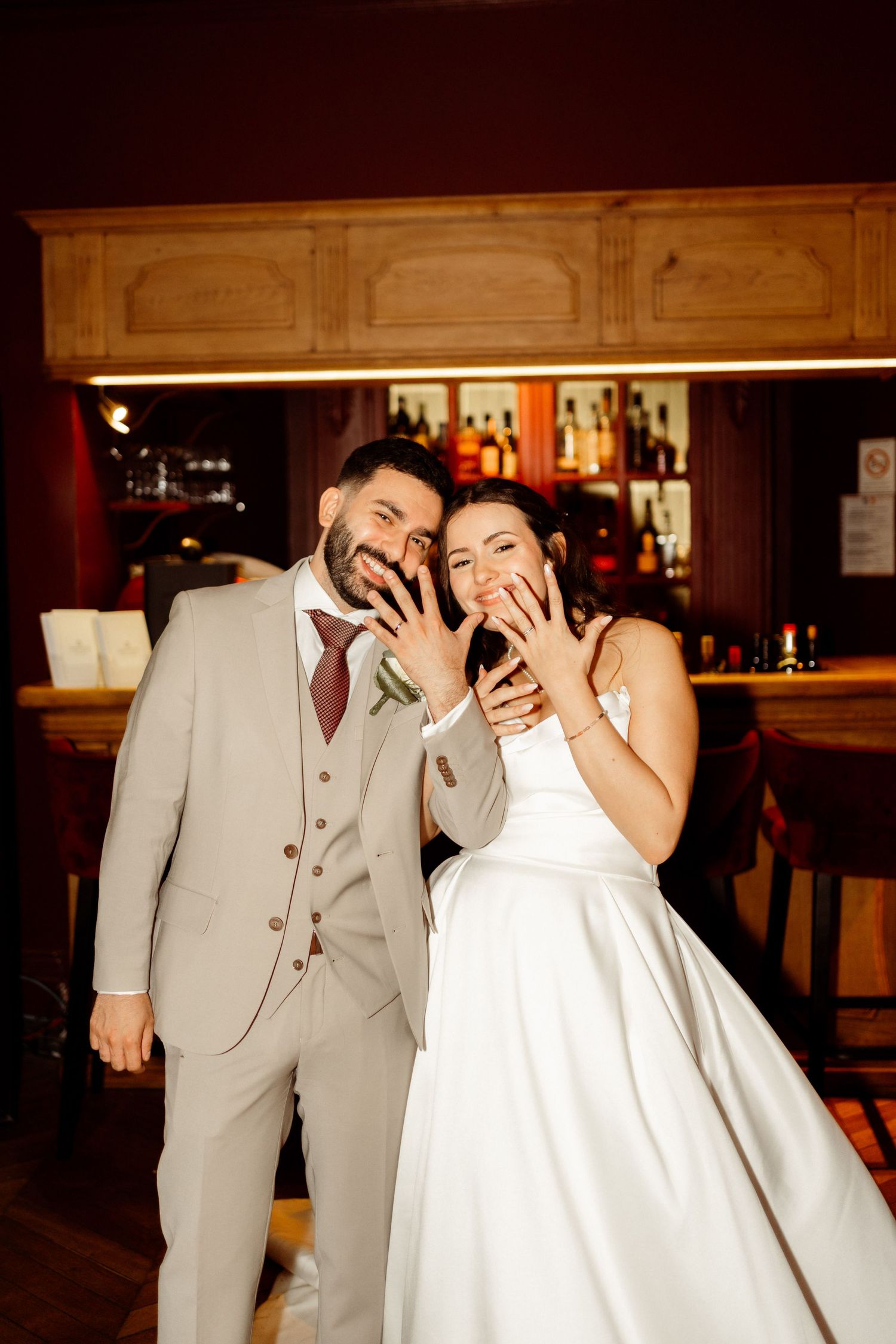 Newlywed couple in formal attire laugh together while showing off rings at a bar reception.