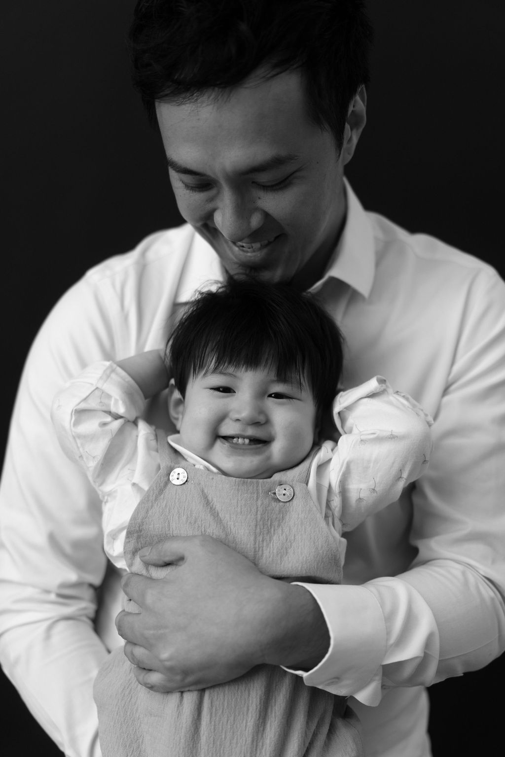 Black and white portrait of a smiling man in white shirt tenderly holding a happy laughing baby girl黑白肖像照，一位身穿白襯衫的微笑男子溫柔地抱著一個快樂大笑的女嬰。