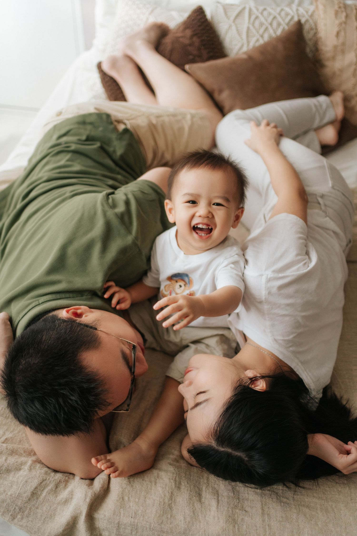 Happy family with parents lying on bed with laughing baby between them in cozy bedroom setting.幸福的一家人，父母躺在床上，中間是咯咯笑的寶寶，溫馨的臥室氛圍。