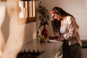 Someone wearing a drapey headscarf writes in a journal at their kitchen counter in warm natural lighting.