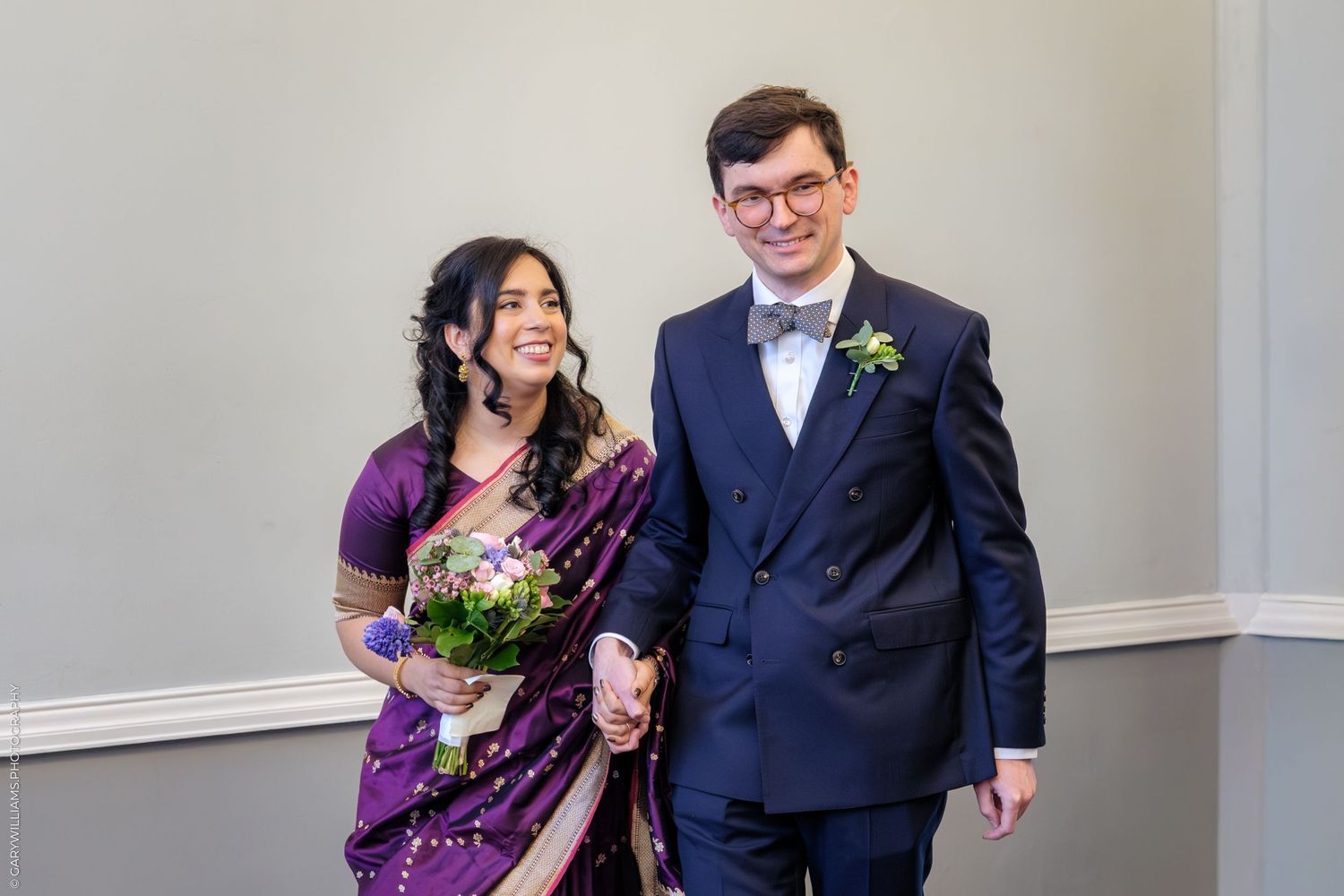 A couple in formal attire stands together at their wedding, with the bride wearing a purple sari.