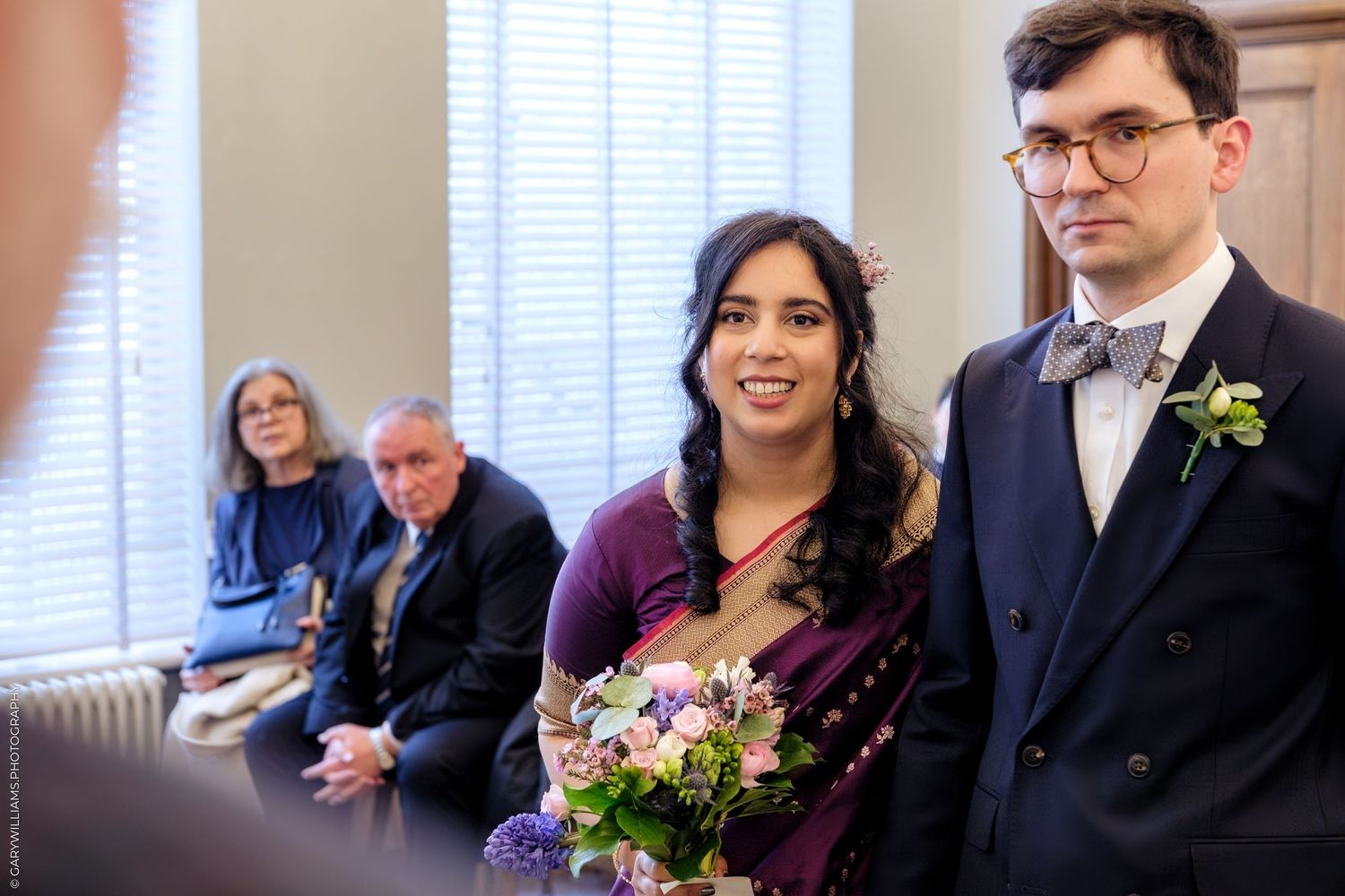 A wedding party stands together in formal attire with bouquets and boutonnières at an indoor venue.