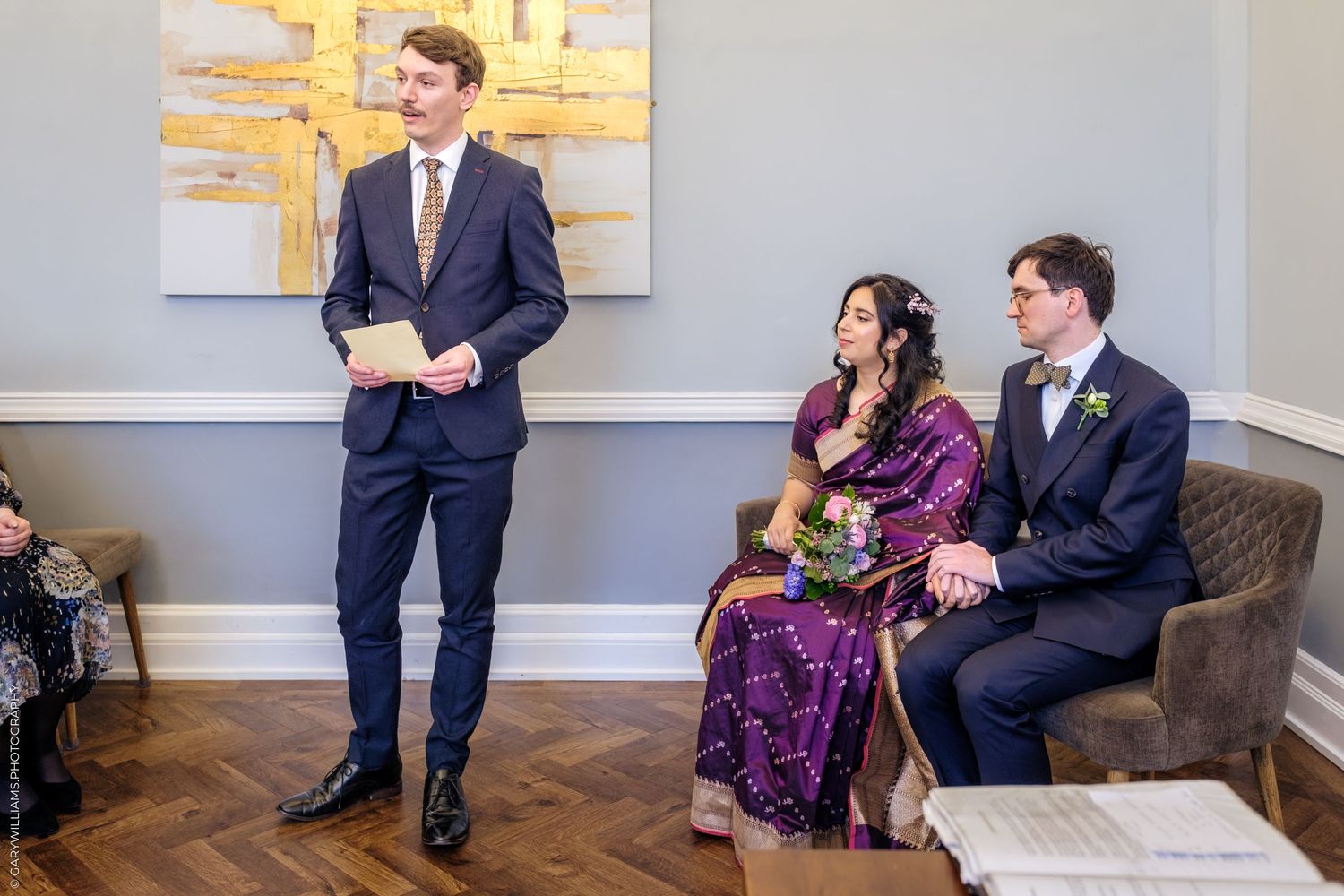 A wedding party sits and stands in a modern room with gray walls and abstract art during a ceremony.