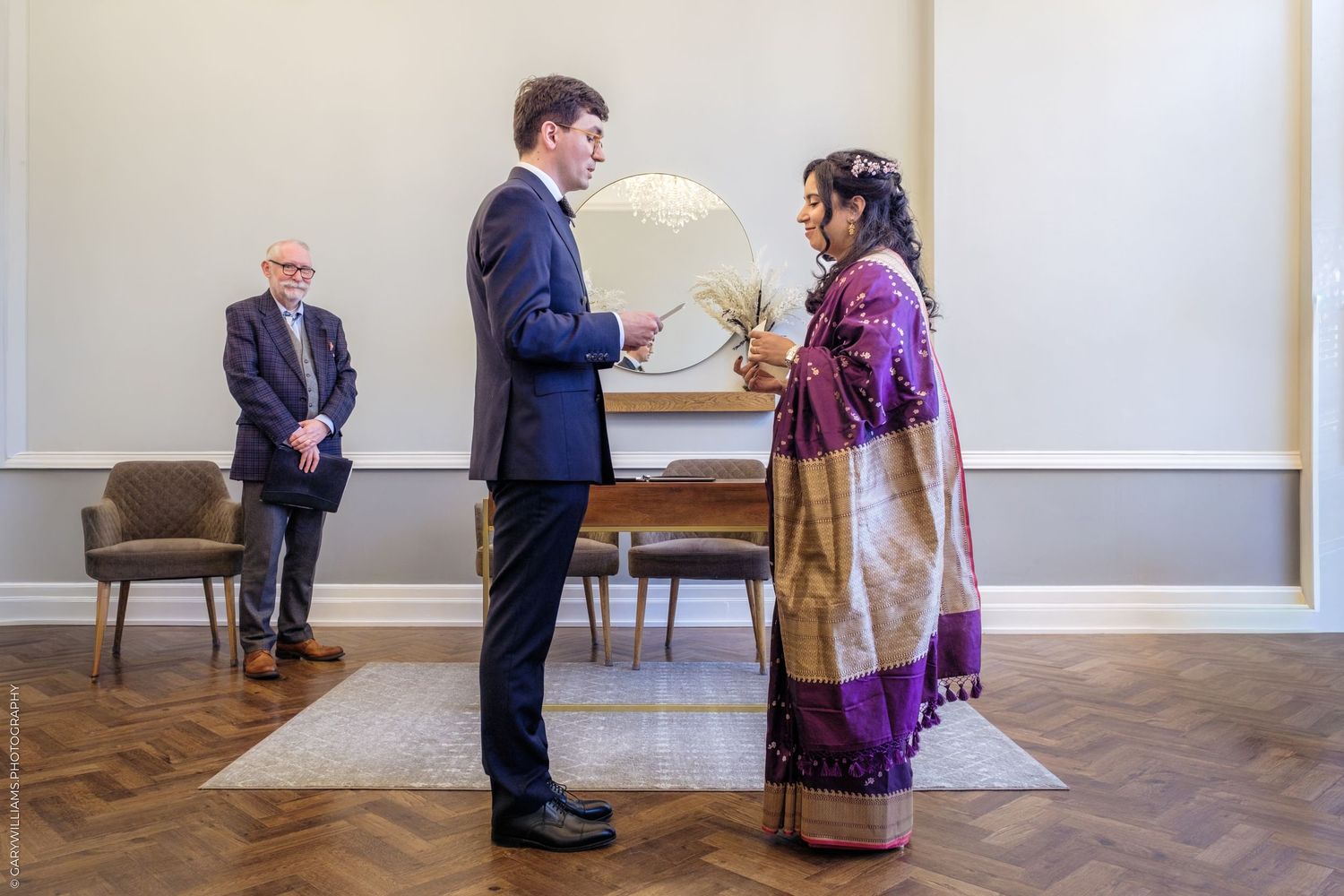 Two people stand in conversation in a minimalist room with white walls and hardwood floors.