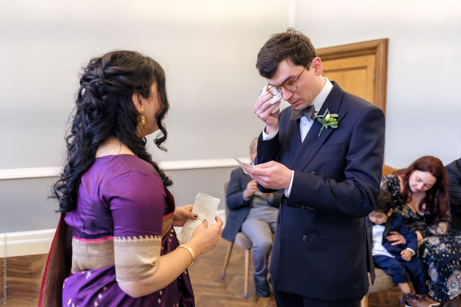 Two people share an emotional moment during a wedding ceremony as they exchange rings in a simple indoor setting.